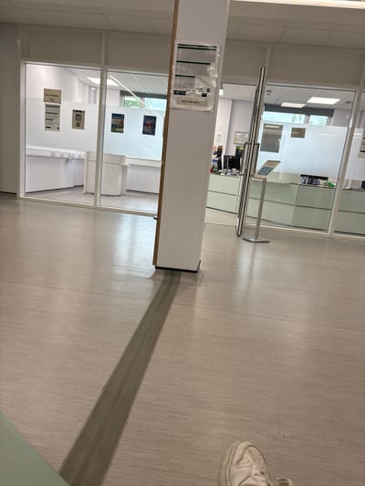 The image shows the interior of a building, possibly a public service facility, in Carlisle, United Kingdom. The perspective is from floor level, angled upwards. The floor is light-colored linoleum or similar material, bisected by a darker, rectangular section, perhaps a guide path. To the left, there are partitioned glass-walled areas, suggesting office spaces or cubicles. Within these areas, some posters or information displays can be seen on the inner walls. A partially visible person's foot in a white trainer is in the upper right. A small kiosk-like structure stands near the glass partition. The building appears to be well-lit with artificial lighting.