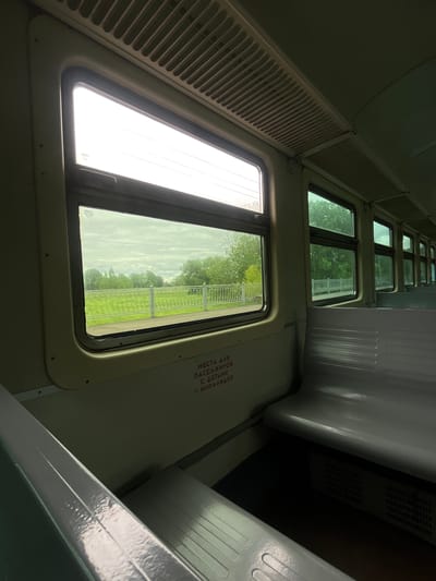 The image shows the interior of a train car, likely in Lampovo, Russia. The view is from inside, looking out through a window. Outside the window, there is a landscape of green fields and trees, possibly a rural area on the outskirts of Lampovo. There are no people visible in the scene. The interior of the train car is mostly white or light-colored, with a series of windows stretching into the distance. There are seats along the side of the train car. There are words in Russian that read: "МЕСТА ДЛЯ ПАССАЖИРОВ С ДЕТЬМИ И ИНВАЛИДОВ". The image is taken at an angle, possibly from someone sitting in a seat.