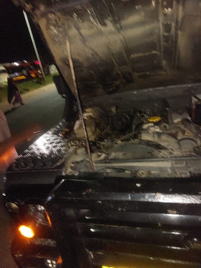 The scene is set at night in Msata, Tanzania. A black utility vehicle, possibly an SUV or truck, is stationary with its hood open, exposing a dirty engine compartment. A yellow cap, likely for oil or coolant, is visible on the engine. The underside of the open hood appears grimy. The front left amber turn signal or hazard light of the vehicle is illuminated. To the left and in the background, a paved road is visible, on which a lone individual, dressed in dark clothing, walks away from the viewer. Further down the road, another vehicle, possibly red or orange, is stopped with its rear lights illuminated. A street light pole stands on the left side of the road, casting light on the environment.