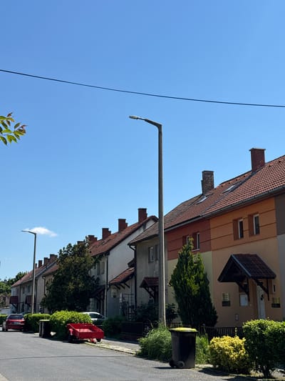 This is an eye-level, outdoor shot of a residential street in Siófok, Hungary. The street is lined with a row of two-story townhouses with red tile roofs and chimneys. Building facades vary in color, from white to beige to orange, and feature dormers. The street is paved and has a sidewalk on the right, along which grow hedges and small trees. A black trash bin is visible near the front of one of the townhouses. Parked cars and a red trailer are visible along the street. Overhead, there is a streetlight with a long arm extending from a pole, and power lines stretch across the blue sky. The sky is clear and bright.