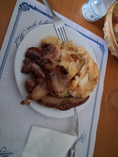 A plate of food, likely a mixed grill with fried potatoes and a fried egg, sits on a white paper placemat with blue decorative borders. The placemat has the text "Enjoy your Meal" printed on it. A fork is visible resting on the plate. To the right of the plate, a basket containing bread and a clear glass with a small amount of liquid can be seen. In the background, a white plastic bottle is also present. The table surface appears to be wood. The overall scene suggests a meal at a restaurant in Mas Aliu, Spain.