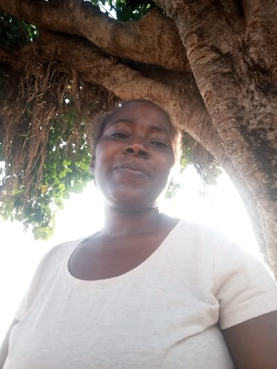 An adult female is depicted in a close-up, upward-angled shot, centered beneath the canopy of a large tree in Gudige Sabo, Nigeria. She has dark skin, short, light-colored hair, and is wearing a light-colored, short-sleeved t-shirt with a round neckline. Her expression is neutral as she looks directly at the camera. Above and behind her, thick, textured tree branches and numerous thin, dark aerial roots or vines are visible, interspersed with green leaves. Bright sunlight emanates from the upper right and behind the subject, casting a bright, slightly overexposed background on the right side of the frame.