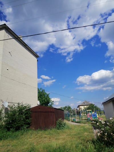 The image depicts a residential area in Lanivtsi, Ukraine, featuring a multi-story brick building on the left, a rusty metal garage, and a playground in the background. The sky is blue with scattered white clouds. The building appears to be an apartment block. The garage is situated in front of the building and is surrounded by overgrown grass. The playground contains colorful equipment, including slides and swings, enclosed by a fence. A residential house is visible behind the playground, and more houses are scattered further in the background. Power lines are strung across the top of the image. The scene is captured on a clear, sunny day.
