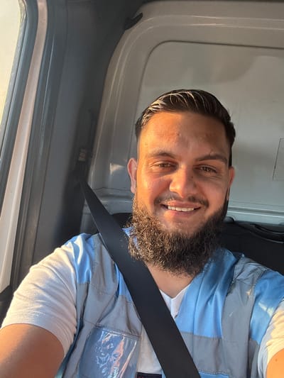 The image shows a man with dark hair and a beard, wearing a blue and gray vest over a white t-shirt, and a black seatbelt. He is smiling and looking at the camera. The setting appears to be the interior of a vehicle, possibly a truck, with the door open on the right side. The lighting suggests it is daytime, with sunlight casting shadows and highlighting his face. The man's expression is cheerful and relaxed. The image is taken from a slightly low angle, looking up at him. There is no visible text in the image. The location context provided is Breendonk, Belgium.