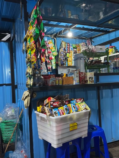 This is a photograph taken in Denasri Kulon, Indonesia, featuring a roadside stall filled with various packaged goods, primarily snacks and drinks. The stall is covered by a blue corrugated metal roof, supported by a metal frame. On the right side of the image, two blue plastic chairs are visible, suggesting a place for customers to sit.

The stall is densely packed with products. In the foreground, several rows of colorful snack bags are displayed, including brands like "Nabati Rebo" and "Vind." To the left, more snack bags are arranged on shelves. In the middle of the stall, a white plastic container is filled with packaged snacks, and next to it, a grey electric appliance, possibly a water dispenser or cooler, is present. Further back, more items are stacked on shelves, including bottles and packaged goods. A single light bulb illuminates the scene.

The photograph appears to be taken during the day, as evidenced by the lighting. The overall impression is of a small, busy local shop operating in a community setting. The clutter and variety of products suggest a convenience store catering to everyday needs. Visible text includes brand names and product descriptions on the packaging, such as "Milkuat," "Oatstik," "Cream," "Kambing Etawa," and "BPA FREE FOOD CONTACT SAFE."