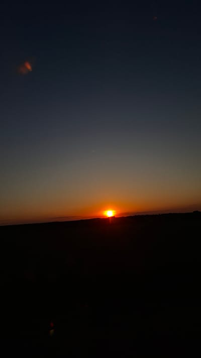 The image shows a horizontal view of a sunset or sunrise. A bright orange sun is visible at the horizon between a lighter-colored sky and a darker area, possibly land or clouds. The lighter sky transitions from gray to orange near the horizon. There are some minor lens flares in the darker areas of the image. The location is Любанскі сельскі Савет, Belarus.