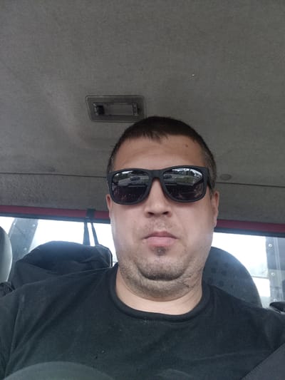A man is taking a selfie inside what appears to be the cabin of a vehicle. He is wearing a black t-shirt and black sunglasses. The interior of the cabin, including the ceiling and a headrest, is visible in the background. The man's face is the primary focus, with a slight smirk. The sunglasses reflect a residential street with houses and trees. The overall lighting suggests it is daytime. The image does not provide any specific clues to pinpoint the location within Čistá, Czechia.