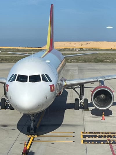 An airplane, likely a Vietnam Airlines aircraft, is parked on an airport tarmac under a clear blue sky. The front of the aircraft is prominently featured, with its white fuselage, cockpit windows, and red and yellow tail fin visible. Two Vietnamese flags, featuring a red background and a yellow star, are affixed to the nose of the plane. The aircraft's engines are also visible, one of which is a red and black jet engine. The tarmac is marked with yellow lines, and a small orange traffic cone is placed near the front landing gear. In the background, a sand dune stretches across the horizon, and a distant structure that appears to be an airport terminal or hangar is visible. The location is identified as Cam Ranh Ward, Vietnam, indicating this scene takes place at Cam Ranh International Airport. No people are clearly visible in the image.