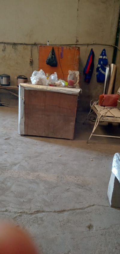 The image shows an indoor scene, possibly a storage room or workshop, in Sumput, Indonesia. The space is characterized by a concrete floor with visible cracks and a background wall. A workbench with a wooden front and white covering the sides stands prominently in the middle. Various objects are placed on the workbench, including containers, bags, and other miscellaneous items. Adjacent to the workbench, there is a metal-framed chair with a red cushion on the seat. The wall behind the workbench has an orange board with a black bag and a purple object hanging on it. Electrical wiring runs along the wall. Two blue and red work coats are also hung on the wall. Other objects are present, including a rice cooker.