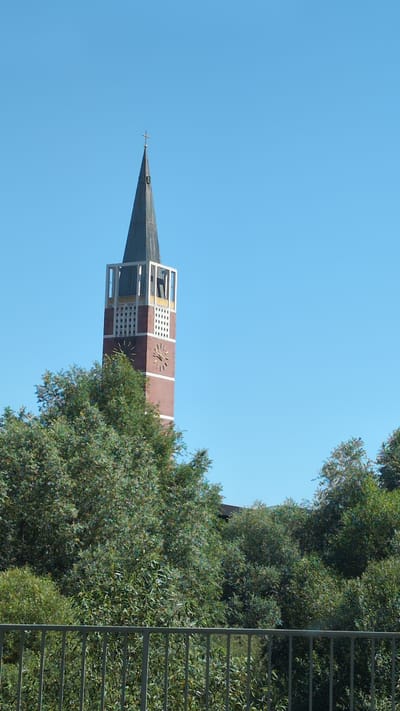 This image, taken on a clear day, shows the tower of the Emmauskirche (Emmaus Church) in Pforzheim, Germany. The tower is tall and slender, with a dark spire topped by a cross. The main body of the tower is constructed of red brick and features a prominent clock face with Roman numerals. Below the clock, there are decorative grid-like elements. Surrounding the base of the tower is lush green foliage, obscuring much of its lower portion. In the foreground, a grey metal fence runs horizontally across the frame. The sky is a uniform, bright blue, with no clouds visible.