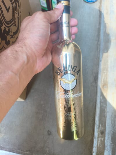 A hand holds a tall, gleaming gold bottle of Beluga vodka. The bottle has a black and gold label with the word "BELUGA" prominently displayed, followed by a circular emblem featuring a golden fish and stylized waves. Below the emblem, intricate lettering reads "Celebration" and a depiction of a tower. The background appears to be an outdoor setting with a gray, textured surface that could be concrete or pavement. There is a brown cardboard box or bag visible to the left of the bottle. The lighting suggests it is daytime, with sunlight casting shadows and reflections on the bottle. The image does not provide any visual information to determine the location in the city of Alchevsk, Ukraine.