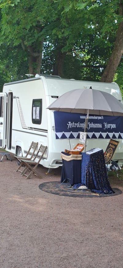 This image depicts an outdoor setup featuring a white caravan, likely serving as a mobile business. In front of the caravan, a table is draped with a dark blue, glittery fabric and covered with a dark blue cloth, on which rests an open suitcase and a book. A large grey umbrella is open and positioned above the table, providing shade. A dark blue banner with white lettering is hung on the caravan, reading "Astrologi Johanna Karpou" and a website address. To the left of the caravan, two wooden folding chairs are placed on the ground. The ground itself is covered in gravel. Behind the caravan, trees with lush green foliage are visible, suggesting a natural or park-like setting. The overall scene appears to be a temporary stall or display for an astrology service, potentially at an event or market. Without further information, the specific location in Turku, Finland, cannot be determined from the image alone.