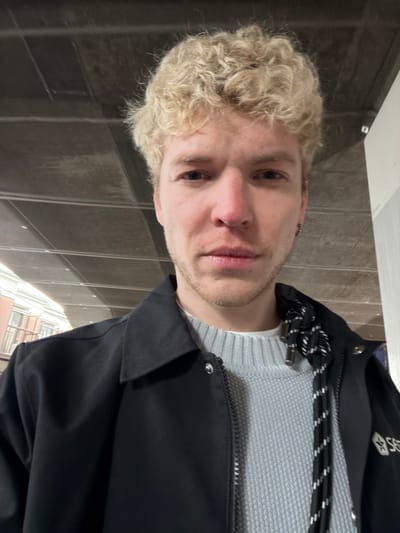 A man with curly, bleached blond hair gazes at the camera. He has fair skin, a faint stubble beard, and wears a light gray sweater beneath a black jacket. The jacket features a distinctive black and white rope drawstring and snap closures. The background includes blurred architectural elements suggesting an indoor or urban environment. The image is rotated, presenting the man's head tilted to the left.
