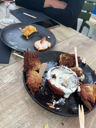 The image shows a close-up view of a wooden table with two black plates of food. The plates contain fried potato fritters, some with a crispy, shredded potato topping, and a small dish of white sauce or dip. One plate has a few pieces of food remaining, while the other has more scattered food remnants. There are wooden skewers inserted into some of the food items. A glass of red wine is visible in the upper left corner, and a person's arm and shoulder, wearing a black top, are in the background. The setting appears to be an outdoor dining area, possibly a restaurant, with a green chair visible in the far background. The lighting suggests it is daytime. The image does not provide any specific information to indicate the location is Saint-Priest-en-Jarez, France.