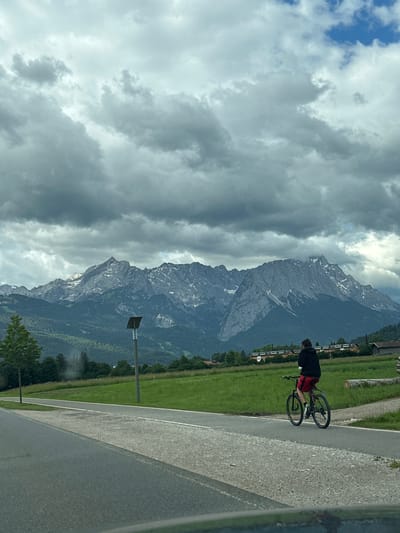 The image depicts a scenic mountain landscape with a cyclist in the foreground. The cyclist, viewed from behind, is wearing a black hoodie and red shorts, riding a dark-colored bicycle on a paved path. The path appears to be a bike path or a side road adjacent to a larger road. Beyond the path, a grassy field stretches towards a line of trees and buildings. In the background, a majestic mountain range dominates the skyline, with its peaks partially covered in snow and shrouded by dramatic, cloudy skies. The presence of a single lamppost in the middle ground suggests an outdoor public area. The scene conveys a sense of outdoor recreation against a stunning natural backdrop. The location is identified as Farchant, Germany.