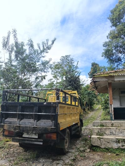A yellow truck with a black cargo bed is parked on a rocky path surrounded by trees. The truck is facing away from the camera, and the license plate is partially visible. To the right of the truck, there is a small, weathered building with a tiled roof and concrete steps. The sky above is blue with scattered clouds. The overall scene suggests a rural or mountainous area.