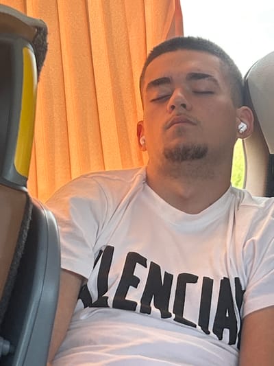 In Diadovo, Bulgaria, a young man with short hair and closed eyes is seen from the chest up, wearing a white t-shirt with black lettering that appears to spell "BALENCIAGA". He has a small goatee and is wearing white wireless earbuds. He is seated, possibly on a bus or in a vehicle, as indicated by the orange curtains in the background and a gray seat armrest in the foreground. The lighting suggests it is daytime.