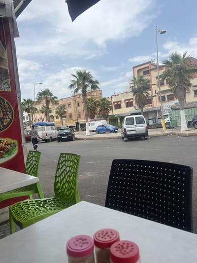 View from an outdoor seating area in Boufakrane, Morocco, including a light-colored table with a black chair, a plastic green chair, and spice shakers, with buildings, palm trees, and parked cars along a street visible in the background.