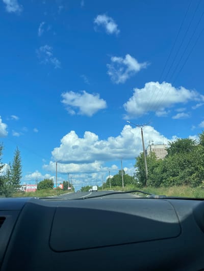 The image is a point-of-view shot from inside a vehicle, looking out at a road on a sunny day. The sky is bright blue with scattered white cumulus clouds. Power lines are visible stretching across the frame. On the right side of the road, there are trees and a multi-story building. On the left side of the road, there are more trees and a billboard. The road curves to the right in the distance, and other vehicles are visible. The dashboard of the vehicle is in the foreground. The scene is consistent with a suburban or semi-rural area.