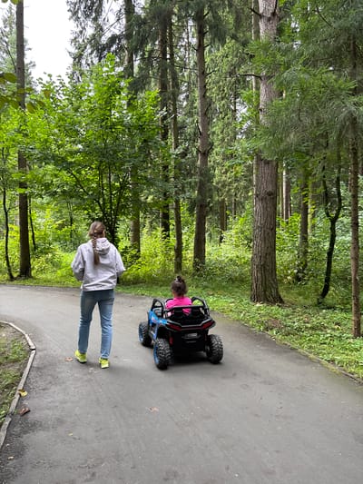 In an outdoor setting with a paved path winding through a dense forest, a teenage girl with braided hair, wearing a light-colored hooded jacket and blue jeans, walks away from the camera. Beside her, a young child sits in a black and blue electric toy UTV. The UTV has large, knobby tires and a roll cage. The child is wearing a bright pink top. The forest is characterized by tall, mature trees with lush green foliage. The path appears damp, suggesting recent rain or dew. The overall impression is of a leisurely outing in a natural park or wooded area.