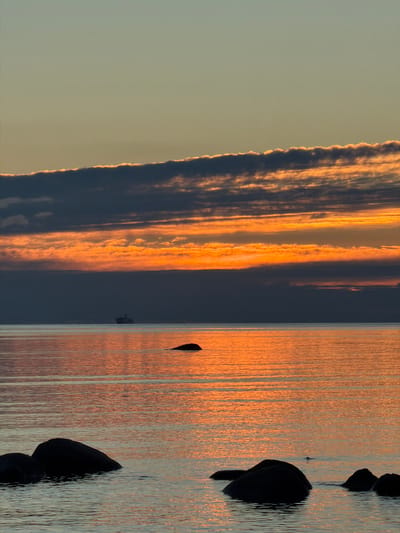 The image depicts a serene seascape at dusk in Haabneeme alevik, Estonia. The sky is a canvas of warm, diffused light, transitioning from a pale gold near the horizon to a soft blue overhead. A band of dark, striated clouds stretches across the middle of the frame, tinged with fiery orange and yellow where they catch the last rays of the sun. Below the clouds, a thin layer of haze softens the distant horizon.
The sea is calm, reflecting the vibrant colors of the sky in shimmering horizontal streaks. A large ship, possibly a ferry or cargo vessel, is visible as a dark silhouette on the water in the distance, almost merging with the haze. In the foreground, several dark, rounded rocks emerge from the water, their shapes softened by the twilight. One solitary rock breaks the surface further out on the water, a small, dark shape against the glowing reflection. The overall impression is one of tranquility and natural beauty as day gives way to night.