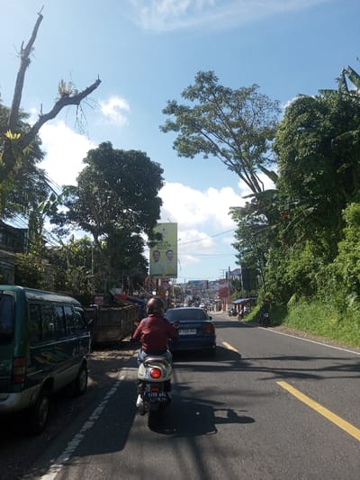 The image depicts a daytime street scene from a moving vehicle's perspective, under a bright blue sky with scattered white clouds. The setting is an urban or semi-urban area in Indonesia, identifiable by the "PARTAI GOLKAR" political billboard and Indonesian vehicle license plates (e.g., "B 3326 NWL" and "F 4788 WAL").

In the foreground, a single individual wearing a dark red jacket, blue jeans, and a light brown full-face helmet is riding a white scooter. The scooter's rear license plate, "F 4788 WAL", is visible, and its red taillight is illuminated. Directly ahead of the scooter is a dark blue sedan, with license plate "B 3326 NWL", also moving forward. The road is asphalt, marked with a dashed white line in the center and a solid yellow line on the right shoulder.

On the left side of the road, a dark green minivan is parked. Behind it, an open-top metal container or skip bin is visible. Further down the street on the left, a large yellow billboard is prominently displayed, featuring two smiling male figures and the text "PARTAI GOLKAR". Overhead power or communication lines crisscross the upper left portion of the frame. Buildings are partially obscured by trees and foliage along the left side.

The right side of the road is characterized by dense, lush tropical vegetation, including tall trees and large-leaved plants resembling banana trees, suggesting a natural embankment or undeveloped area. Additional vehicles, including cars and motorcycles, are visible in the distance, indicating ongoing traffic. Sunlight is strong, casting prominent shadows to the right and front of the moving vehicles.