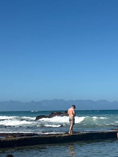 The image shows a view of the beach in Muizenberg, South Africa, with a clear blue sky taking up most of the frame. The coastline is visible on the right side, with waves crashing against the rocks. A man in shorts stands on the rocks, near the water. In the background, there are hazy mountains visible.