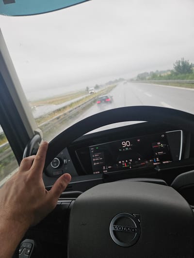 The image shows a first-person view from the driver's seat of a Volvo truck, as indicated by the Volvo logo on the steering wheel. The driver's left hand is visible, resting on the steering wheel. The dashboard displays a speed of 90 km/h and other vehicle information. Outside the vehicle, it is raining, and the road appears wet. Several vehicles are visible on the highway in front of the truck. The location is identified as Stephansposching, Germany, likely on a highway. The overall scene depicts a rainy day drive on a highway.