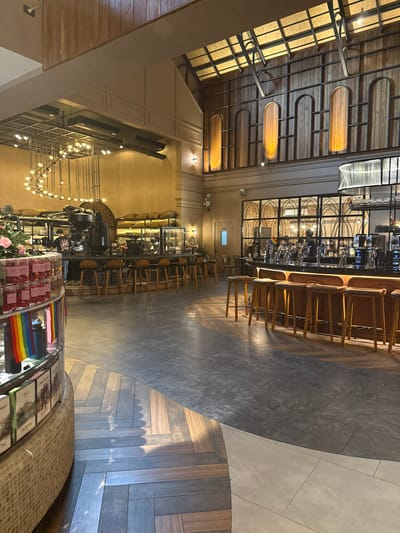 The image depicts the interior of a modern, stylish restaurant or bar with a warm and inviting atmosphere. The scene is shot from a high angle, looking down and across the space.
The dominant elements of the scene are the polished wooden ceiling with embedded lighting, the sleek bar area with numerous stools, and the varied flooring, which transitions from dark, textured tiles to lighter, matte tiles and then to a herringbone pattern of wooden planks. The bar is well-lit, showcasing rows of bottles and glasses. Several people are visible behind the bar, engaged in preparing drinks or serving customers. The seating area in front of the bar has multiple wooden stools.
In the foreground, there's a display case filled with small, individually packaged items, possibly desserts or treats, and a prominent display of rainbow flags, suggesting a Pride month celebration or an inclusive establishment. Behind this, a large, dark coffee machine is visible, hinting at a cafe or a place that serves coffee. Further back, a decorative chandelier with many small lights hangs, adding to the ambiance. The walls are a warm, neutral color, and the overall lighting suggests it is either evening or the interior lighting is designed to create a cozy atmosphere.
The location context of Ban Na Chom Thian, Thailand, is not explicitly visible in the image, but the style and presentation are consistent with a contemporary establishment found in such a tourist-oriented area. There is no visible text in the image that is legible or provides specific information.
