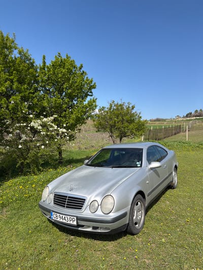 The image shows a silver Mercedes-Benz CLK coupe parked on a grassy field. The license plate number is CB 9443 PP. A tree with white blossoms is to the left of the car, and a vineyard is visible in the background. The sky is clear and blue. The location is Djigurovo, Bulgaria, based on the license plate designation. The car appears stationary. There are no people visible in the scene.