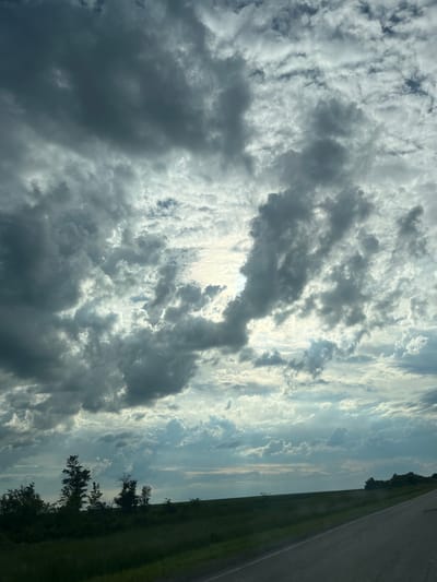 The image depicts a dramatic sky with heavy, dark, and light grey clouds, suggesting an approaching storm or post-storm conditions. Sunlight is breaking through in patches, creating highlights and casting shadows. Below the turbulent sky, a low, green landscape stretches out, lined with a dark silhouette of trees and vegetation along a distant horizon. A paved road is visible in the foreground, angling from the bottom right corner of the frame towards the horizon, indicating a journey through a rural or semi-rural area. The scene is devoid of any people or specific structures, focusing solely on the natural environment and atmospheric conditions. The location is identified as Ozerny Aul District, Kazakhstan.