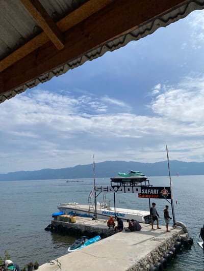 The image depicts a waterfront scene at Tuk Tuk, Indonesia. A concrete pier extends into a calm body of water, with several people present on it. On the pier, a group of individuals are gathered near a boat that has "PARASAILING" written on its side. Another group of people are standing further down the pier. A sign with "SENODO SAFARI SAMOSIR" is visible.
Two jet skis are present; one is parked on the pier and another is in the water. In the background, a range of verdant, sloping hills forms the horizon, with a hazy sky overhead dotted with clouds. A portion of a corrugated roof structure is visible in the foreground, casting a shadow over part of the scene.