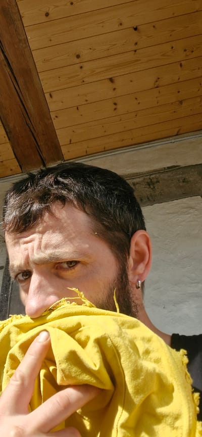 A close-up, vertical, outdoor portrait shows a man's face and upper body, partially obscured by a bright yellow cloth. The man has short, dark hair and a beard, with a worried expression on his face as he squints. He is wearing a silver hoop earring in his left ear. The background consists of a wooden ceiling and a rough, light-colored wall. The image appears to have been taken in Wattenwil, Switzerland, suggesting a domestic or outdoor setting in that location. The lighting indicates it is daytime.