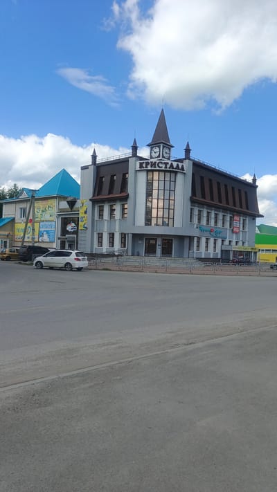 This is a medium-shot, eye-level, outdoor, full-color photograph taken on a sunny day with scattered clouds. The scene depicts a street in Kuybyshev, Russia, with a modern, multi-story building dominating the right side of the frame. The building has a dark, angular roof and a light gray facade with large, reflective windows. A prominent sign with the Cyrillic text "КРИСТАЛЛ" (Kristall) is visible on the upper portion of the building, along with two clock faces.

To the left of the main building are other businesses, one with a bright blue, pitched roof, and another with a wooden facade and awnings. Cars are parked along the street in front of these businesses, including a white minivan, a black sedan, and two smaller yellow cars. A metal fence runs along the sidewalk in front of the buildings. The street in the foreground appears to be made of asphalt, with some wear and tear. The overall impression is that of a commercial area on a bright, clear day. The time of day appears to be daytime, likely mid-morning or afternoon, given the bright sunlight and shadows.