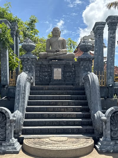 The image displays an ornate stone religious structure, likely a temple or shrine entrance, bathed in bright daylight under a clear blue sky dotted with scattered white clouds. The central focus is a light-colored stone statue of Buddha, seated in a contemplative Dhyana Mudra pose with hands resting in his lap, positioned atop a dark gray, heavily ornamented stone pedestal.
A grand set of dark gray stone stairs leads upwards towards the statue, with each step meticulously carved with repetitive floral or geometric patterns. The stairway and statue are framed by robust, dark gray stone pillars featuring elaborate, multi-layered capitals and bases, all adorned with detailed relief carvings. Complementing these are ornate balustrades or railings, also crafted from dark gray stone, which incorporate decorative urn-shaped finials and swirling motifs.
Lush green trees and foliage flank the stone architecture, providing a vibrant natural backdrop. In the mid-ground, parts of a building with a traditional terracotta-tiled roof are visible, alongside a section of gold-colored ornamental metalwork, possibly from a gate or fence.
A rectangular plaque, light in color and containing unreadable text, is prominently displayed on the front of the dark gray pedestal beneath the Buddha. The strong illumination and distinct shadows suggest a sunny midday or early afternoon setting. This serene and culturally rich scene is located in Matara, Sri Lanka.
