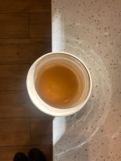 A top-down, slightly oblique view captures a single white disposable-style cup containing a light brown liquid, consistent with coffee or tea, resting on a speckled light-colored countertop. The countertop occupies the right half of the frame and displays distinct wavy light patterns, suggesting filtered sunlight. The left half of the frame features a dark brown wooden floor composed of rectangular planks. The toes of two dark-colored shoes are partially visible at the bottom left, positioned on the wooden floor. The scene is located in San Antonio de Padua, Argentina.
