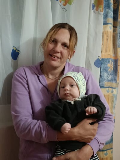 A woman is holding a baby in front of a light-colored curtain with a repeating pattern of blue and yellow floral motifs. The woman has shoulder-length blonde hair and is wearing a light purple hooded sweatshirt. She is smiling and looking at the camera. The baby is wearing a light green bonnet with a pattern and a dark gray long-sleeved shirt with white trim. The baby is looking at the camera with a neutral expression. The background appears to be an indoor setting, possibly a home.