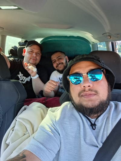 Three men are in the back of a car, appearing comfortable and relaxed. The man in the foreground is wearing blue mirrored sunglasses and a gray hoodie, with a seatbelt visible across his chest. Behind him, another man smiles, wearing a white t-shirt. A third man, wearing a black t-shirt, is positioned further back, and has a bandage on his hand. The location cannot be determined from the image with certainty.