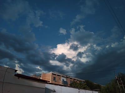 The image, taken in Karshi City, Uzbekistan, depicts a twilight sky filled with dramatic, dark, and some luminous clouds. In the foreground, the top portion of a white building with a red-tiled roof is visible on the left. To the right, a multi-story building with an orange facade and numerous windows is partially obscured by trees. Power lines are visible stretching across the sky. The overall scene conveys a sense of impending weather change.