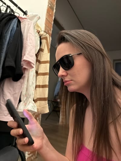 A person with long brown hair and sunglasses is looking down at a phone. The phone is in their right hand. The person is wearing a pink top. A pile of folded laundry sits behind the person. The laundry consists of beige striped fabric, pink fabric with a floral pattern, a solid light pink fabric, and a dark-colored fabric. A brick wall is visible behind the laundry.