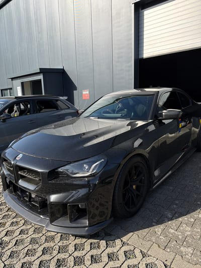 The scene is set outdoors in Meuspath, Germany, specifically in a paved area adjacent to a modern, dark gray industrial-style building. In the foreground, a black BMW M2 coupe is parked, facing slightly left from the viewer. It features an aftermarket carbon fiber front splitter with integrated aerodynamic elements, black multi-spoke wheels, and Michelin performance tires. The vehicle's surface shows a fine layer of dust. Colored vertical stripes (red, yellow, blue) are affixed to the passenger side mirror and front door. The headlights are illuminated. Visible through the front windshield is a racing bucket seat with a "C" logo on the headrest and a red accent on the steering wheel.
Behind the BMW, to the left, a dark gray performance hatchback or wagon is parked. This vehicle features a roof spoiler and an internal roll cage, visible through the rear passenger window. White racing bucket seats are discernible through the driver's side window. The number "12" is visible on a smaller, dark gray structure with a window, positioned to the left of the main building. To the right, a large white roll-up garage door is partially open, revealing a dark interior. The ground is paved with interlocking concrete pavers in a patterned arrangement. The area is illuminated by direct sunlight, casting clear shadows from the vehicles and structures. No individuals are present or actively engaged with the vehicles.