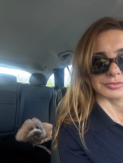 The image displays the interior of a private vehicle, with the specific location within Alcobaça, Portugal, not discernible from the visual evidence. The foreground is dominated by the partial view of an adult female, positioned as if capturing a self-portrait. She has light brown hair, wears dark tortoiseshell-framed sunglasses, and a dark blue collared top. A gold chain is visible on her neck. Her lips are slightly parted, revealing a light pink color. In the background, a small, light brown, curly-haired dog is resting on a dark textile covering the rear passenger seat. The dog's mouth is open, exposing its teeth. The vehicle's interior features dark-colored rear seats with headrests and a light grey headliner. Daylight is visible through the rear windows, suggesting an outdoor environment beyond the vehicle.