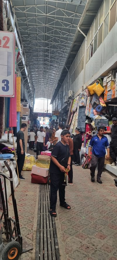 The image depicts an indoor market scene in Bokhtar, Tajikistan. The market appears to be a covered bazaar with a metallic, arched roof structure overhead. The narrow walkway is lined with stalls displaying various goods, including textiles, bedding, and boxed items. People are seen walking through the market, some carrying shopping bags and others interacting with vendors.
In the foreground, a young boy in a dark blue t-shirt and pants is holding a golden trophy. To his left, a trolley with wheels is parked. Further down the walkway, several individuals are walking. On the right side, a man in a blue athletic jacket is walking with a box. The ground is paved with decorative tiles, and a metal grate runs along the walkway. The overall atmosphere suggests a bustling commercial environment.