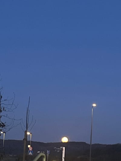The image presents a scene at dusk or dawn in or near Mensdorf, Luxembourg, characterized by a predominantly blue sky. A roadside utility pole stands on the right side of the frame, partially obscured by branches. Traffic signs are attached to the pole, one of which is circular, with the number "30" visible. Two streetlights are visible in the sky; the one on the bottom is bright. A dark, undefined mass, potentially representing a tree-covered area or a hill, forms the background on the right edge.