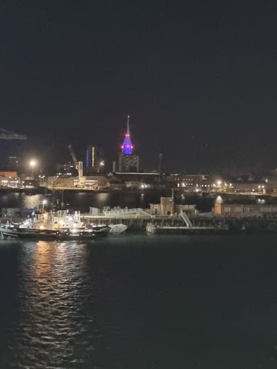 This image depicts a nighttime scene of Portsmouth Harbour, United Kingdom. In the foreground, a body of dark water reflects the lights of the city. A tugboat is visible on the water, illuminated by its own lights and the lights from the shore. In the middle ground, the Portsmouth waterfront is lined with various buildings and industrial structures, including cranes. Dominating the background skyline is the iconic Spinnaker Tower, lit up with purple and blue lights. The overall impression is one of an active port city at night.