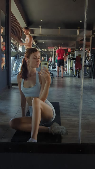 Here's a forensic description of the image:

**Location:** Gym in Pererenan, Bali, Indonesia.

**People:**
*   A woman (presumably taking the photo), wearing a light blue sports bra and light grey shorts, with white sneakers and socks, holding a cell phone.
*   In the background, at least three other individuals: one shirtless man working out on a machine, one man in a red shirt and black shorts, and one unidentified person at a treadmill in the lower left corner of the reflection.

**Objects:**
*   Gym equipment (various weight machines, treadmills).
*   A black yoga mat.
*   A cell phone (presumed to be the phone taking the photo).
*   A mirror reflecting the gym environment.

**Actions:**
*   The woman is taking a selfie in a gym mirror.
*   Other individuals are working out in the gym.

**Environment:**
*   A modern gym with dark ceilings and light-colored tile or laminate flooring.
*   The gym has standard fitness equipment arranged in rows.

**Interactions:**
*   The woman is interacting with her cell phone to capture the selfie.
*   The other individuals in the gym are working out individually.