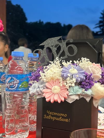 The image captures a detailed view of a graduation celebration, specified to be in Kaspichan, Bulgaria. In the immediate foreground, a black hexagonal box serves as a centerpiece, containing an arrangement of artificial flowers. These flowers are varied in color, including light pink, lavender-purple, cream-white, and light blue/teal, and are interspersed with white baby's breath, suggesting a decorative bouquet, possibly composed of soap or wax. A silver glittery cutout displaying the numbers "2026" and a graduation cap sits prominently at the back of the box, indicating the year of graduation. Gold Cyrillic text, "Честито завършване!", meaning "Happy Graduation!", is embossed on the front face of the box.

To the left of the centerpiece, multiple clear plastic water bottles with bright blue caps are visible. The label of the closest bottle clearly shows the brand name "Велена" (Velena) and "трапезна вода" (table water), accompanied by a logo featuring a water droplet and a green leaf. These bottles rest on a red surface, likely a tablecloth. A segment of a clear drinking glass is visible in the bottom right corner of the frame.

In the midground, several individuals are partially visible behind the table setup. On the far left, a portion of a person's head with dark hair is seen. Further back, the upper torso and back of a person wearing a white shirt are visible, facing away from the camera. To the right of the center, another individual's back and brown hair are discernible, also facing away. The background consists of a dark blue sky, characteristic of dusk or nighttime, with the indistinct silhouettes of trees and dense foliage against it, suggesting an outdoor or semi-outdoor setting for the event.