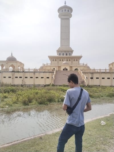 The photograph shows a person standing with their back to the camera, observing a large building across a body of water. The subject is a male with dark, curly hair, wearing a light blue t-shirt, dark blue jeans, and a black sling bag across his shoulder. He is looking down at something in his hands, possibly a mobile device.

In the mid-ground, a tranquil body of water, likely a river or canal, stretches horizontally, separating the person from the background structure. The banks of the water are covered with green grass and dense vegetation.

The background is dominated by a magnificent, light-colored building complex, strongly resembling a mosque or an Islamic cultural center, in Kruengmate, Indonesia. The architecture features a prominent, very tall minaret with a multi-tiered, circular top section and a small dome. The main building includes multiple smaller domes, ornate railings, and a wide set of stairs leading to an elevated entrance. The lower sections of the building, particularly the wall facing the water, show rectangular openings and some signs of wear or construction activity. The sky is bright and overcast, suggesting daytime with diffused sunlight. No text is visible in the image. The overall scene depicts a serene and observational moment against an impressive architectural backdrop.