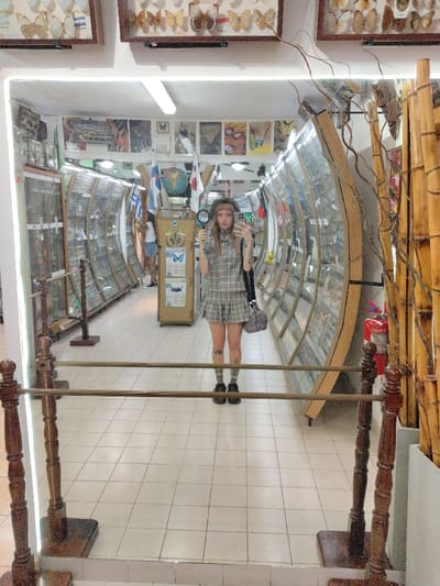 The image captures a mirror selfie taken indoors, likely within a natural history museum, insectarium, or a similar exhibition space dedicated to butterflies and insects.
In the center of the mirror's reflection stands a person taking the selfie. This individual has long, light-colored hair with bangs, and is wearing a dark beret or hat. They are dressed in a grey and white plaid collared shirt and a matching pleated mini skirt, along with plaid knee-high socks and black shoes. Visible tattoos adorn their legs and left arm. In their right hand, they hold a magnifying glass, while their left hand holds the phone used to capture the image. Their expression is neutral.
The background reflected in the mirror reveals a long, narrow room with a high ceiling. On both the left and right sides, there are long, curved display cases, resembling tunnels, filled with numerous specimens, most likely insects or butterflies. These cases are made of glass and wood. On the far left, another person is faintly visible further down the aisle. On the far right, behind the curved display case, a red fire extinguisher is mounted on the wall, and several tall bamboo stalks are placed vertically against the wall.
In the central background, between the curved display cases, a tall wooden display stand is visible. On top of this stand is a small globe. Below the globe, there are framed exhibits, possibly of butterflies. A blue poster or sign with a large butterfly image and text (partially visible, including "BUTTERFLIES", "Papilio memnon", and "Papilio") is attached to the stand. Above this central display, various flags are hanging, including what appears to be a Japanese flag and possibly a Greek or Israeli flag. The walls above the display cases are adorned with multiple framed pictures or posters featuring artistic representations or close-ups of insects.
The ceiling of the room is white, with long fluorescent strip lights providing illumination. The floor is covered with light-colored square tiles.
In the immediate foreground, a wooden railing with turned posts and horizontal wooden bars runs across the bottom of the mirror, separating the viewer from the reflection. Above the mirror, on the wall, several framed exhibits of butterflies are visible, consistent with the theme of the reflected room.