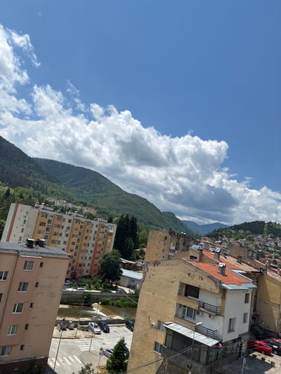 The image depicts a view of Vlahovo, Bulgaria, featuring a cityscape with apartment buildings nestled against a backdrop of lush, tree-covered mountains. The sky above is a vibrant blue, dotted with fluffy white clouds. In the foreground, a tall apartment building with a beige and yellow facade is visible, featuring numerous windows and balconies. The building on the far left has the Bulgarian flag colors (white, green, and red) painted vertically on its side.
Below the buildings, a small river flows, with a concrete embankment and a pedestrian crossing visible on the street. Several cars are parked along the street and near the river. The architecture of the buildings suggests a blend of older and more modern construction. The overall scene conveys a sense of a moderately sized town or city situated in a mountainous region.